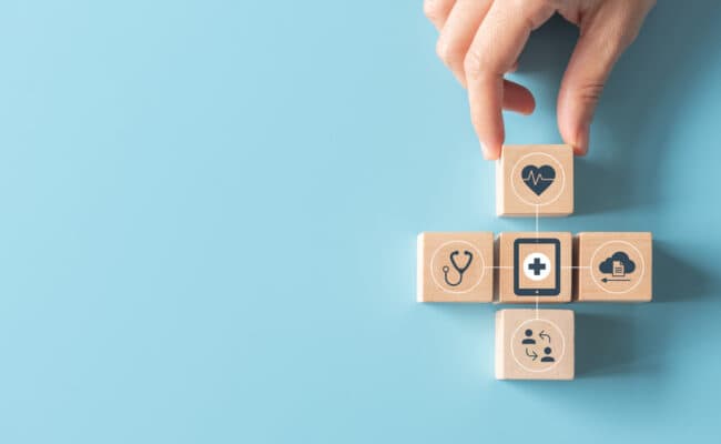 Eine Hand arrangiert fünf Holzwürfel mit verschiedenen Healthcare-Symbolen, die den Connected-Healthcare-Ansatz von avodaq symbolisieren